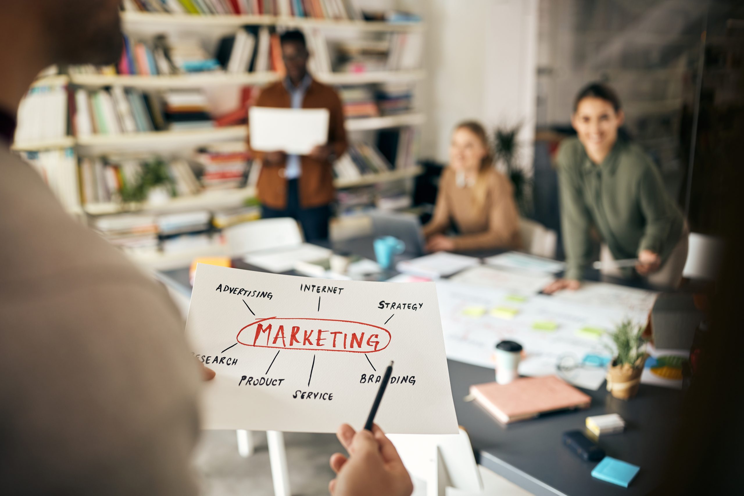 Foglio con MARKETING in rosso e mappa concettuale, sullo sfondo colleghi attorno a un tavolo pieno di post-it